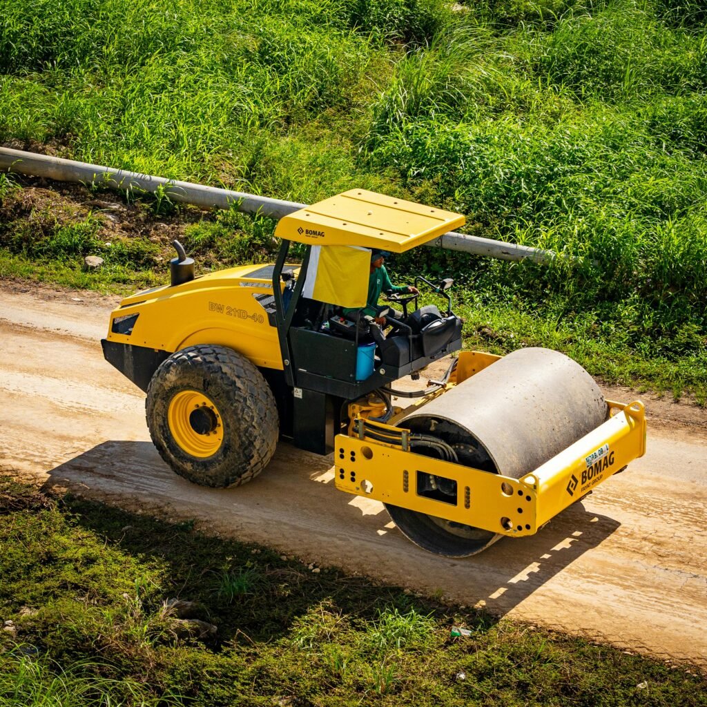 Aerial shot of a yellow road roller on a rural dirt path surrounded by green grass.